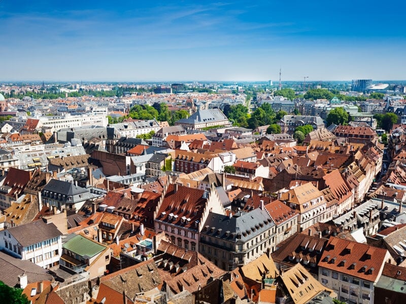 Vue aérienne du centre d'une ville européenne aux bâtiments historiques, avec des toits marron et des rues étroites, contrastant avec des zones urbaines modernes en arrière-plan sous un ciel bleu déga