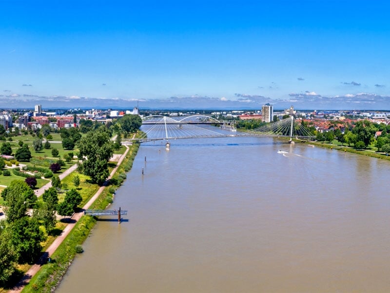 Vue aérienne d'un fleuve traversant une ville, avec un pont suspendu moderne et des rives aménagées de parcs et d'allées arborées. Ciel clair avec des nuages épars.