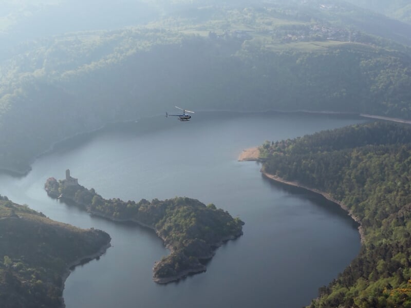 Hélicoptère en vol au-dessus d'un lac entouré de collines boisées, avec une petite île et une tour en ruines.