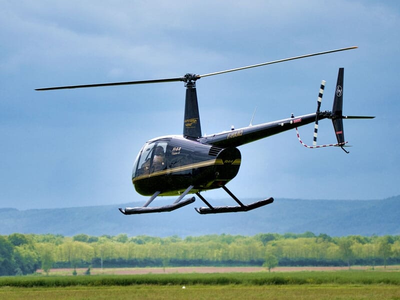 Hélicoptère noir en vol au-dessus d'une prairie, avec des arbres et des montagnes à l'horizon.