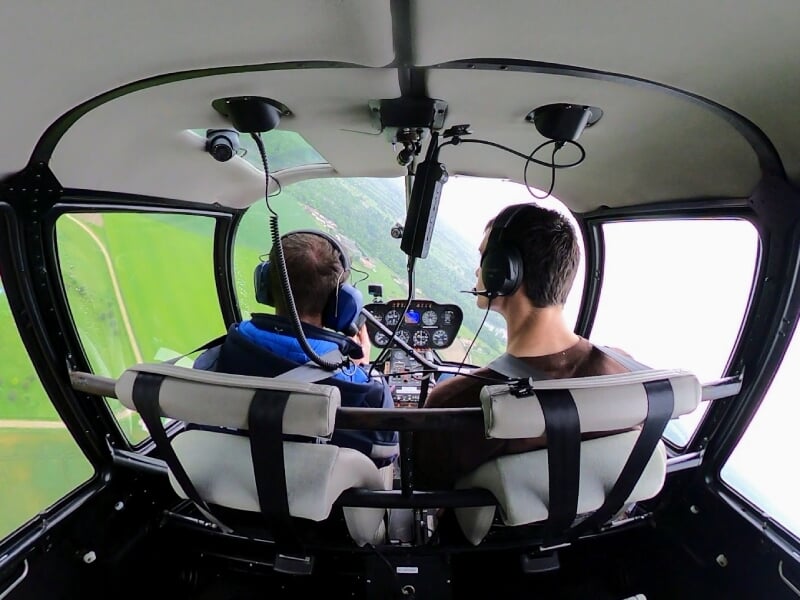 Vue arrière de deux hommes dans un hélicoptère en vol, survolant une campagne verdoyante, avec des instruments de vol visibles.