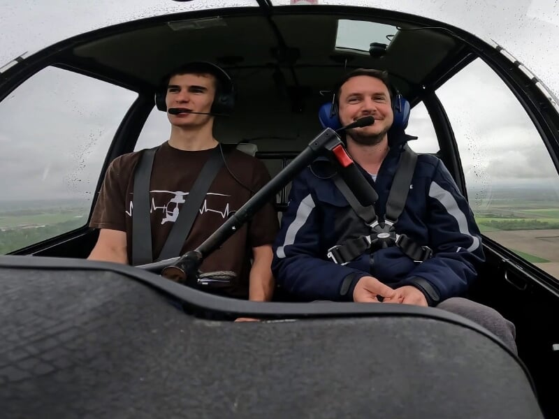 Deux hommes assis côte à côte dans un hélicoptère en vol, portant des casques audio et souriant, avec une vue extérieure nuageuse.