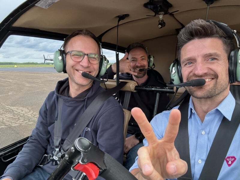 Trois hommes souriants avec des casques audio dans un hélicoptère, l'un faisant le signe de la paix et un autre faisant un geste de téléphone.