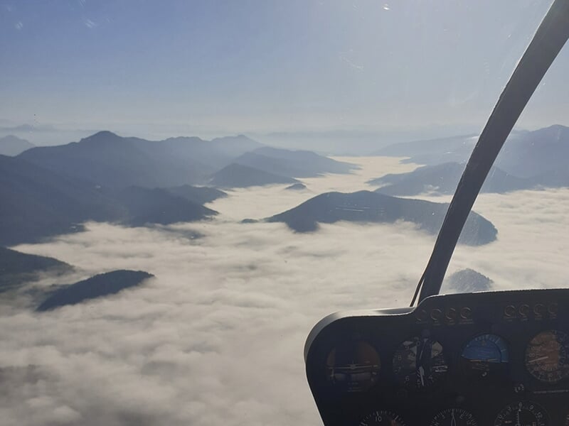 Vue depuis le cockpit d’un hélicoptère survolant une mer de nuages et des montagnes à l’horizon.