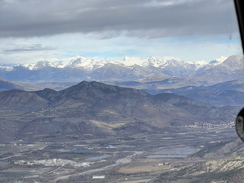 Panorama de montagnes enneigées et vallées verdoyantes capturé depuis un hélicoptère.