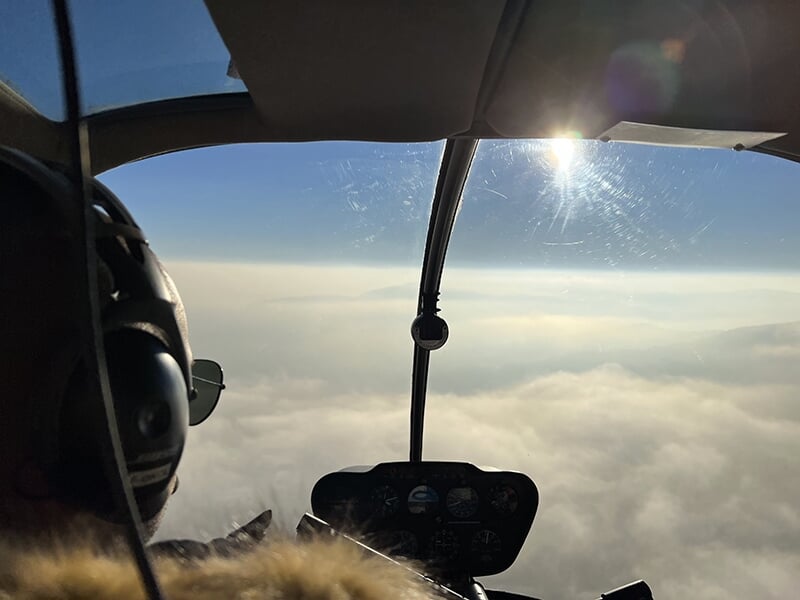 Lever du soleil vu depuis un cockpit d’hélicoptère, avec une mer de nuages à l’horizon.
