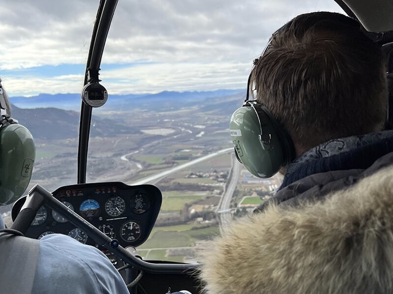 Vue depuis l’intérieur d’un hélicoptère en vol, avec des paysages et des routes visibles en contrebas.