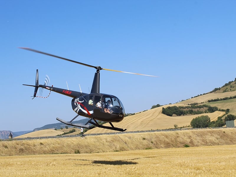 Hélicoptère noir volant au-dessus d’un champ doré, avec des collines en arrière-plan.