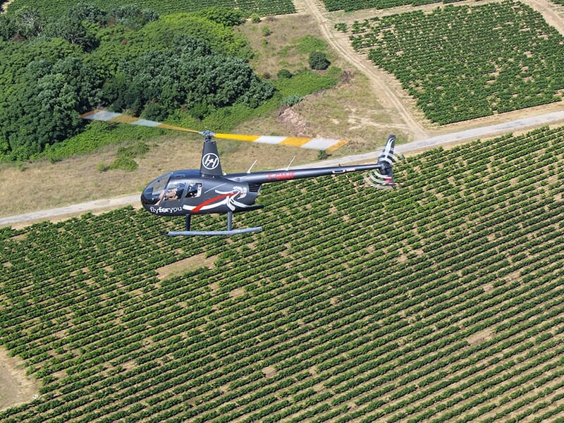 Hélicoptère noir en vol au-dessus de vastes champs de vignes vus d’en haut.