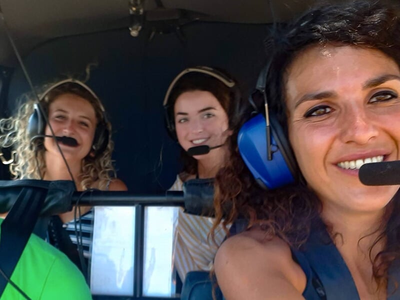 Trois passagères souriantes à l’intérieur d’un hélicoptère, portant des casques avec microphones.