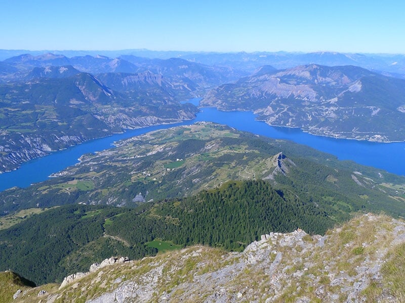 Vue aérienne panoramique d’un lac bleu profond entouré de montagnes et de vallées verdoyantes.
