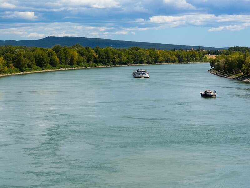 Large rivière calme avec des bateaux de plaisance, bordée de forêts et de collines à l'horizon.