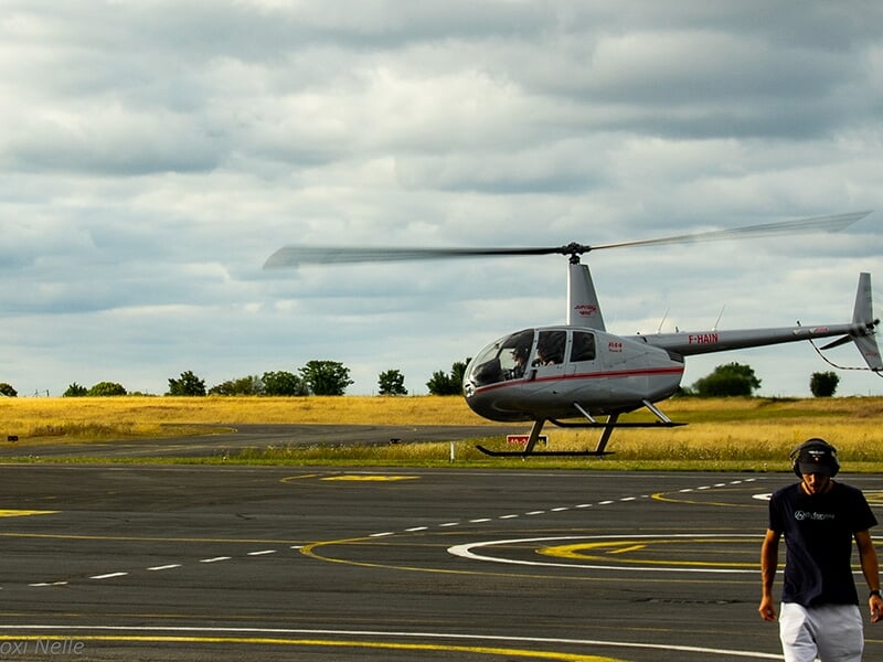 Hélicoptère en vol stationnaire sur une piste d'atterrissage, ciel nuageux et paysage rural en arrière-plan.