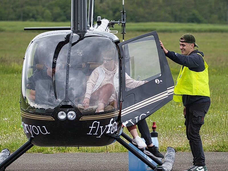 Passager montant à bord d’un hélicoptère noir, assisté par un membre de l’équipe au sol portant une chasuble jaune.