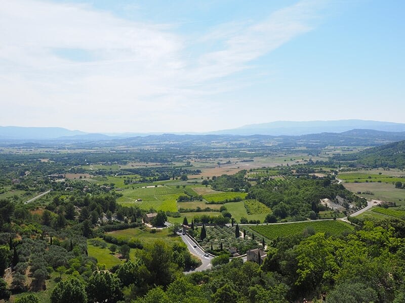 Vue panoramique sur les vastes champs et vignobles de la région du Luberon, avec des routes sinueuses et des collines à l'horizon.