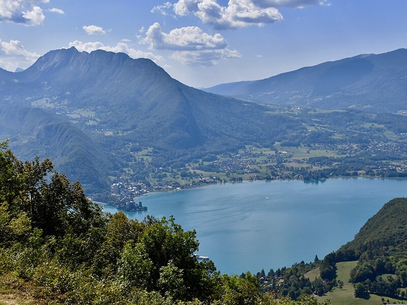 Vue panoramique du lac d’Annecy entouré de montagnes verdoyantes sous un ciel légèrement nuageux.