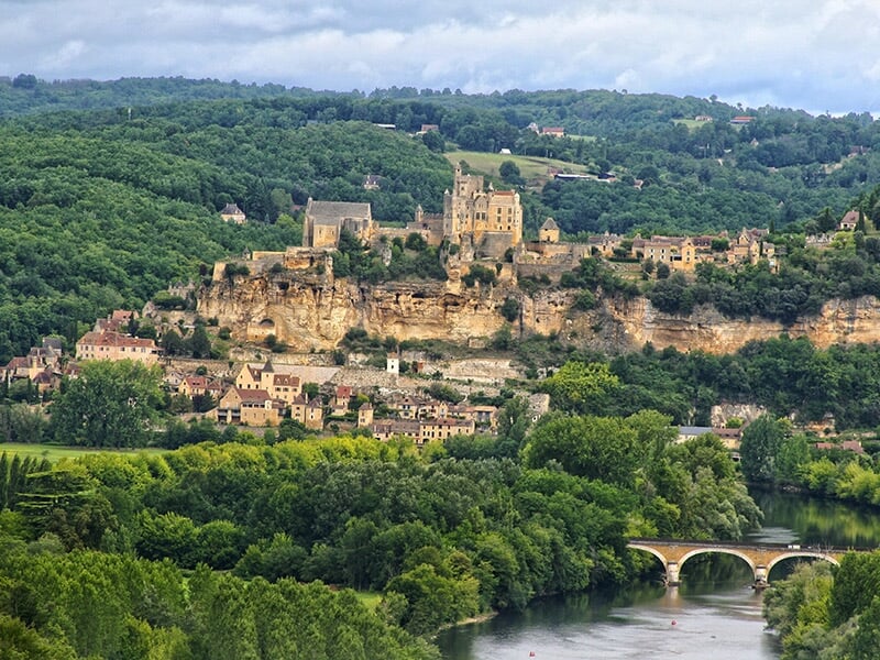 Pont en pierre traversant la rivière Dordogne, encadré par des champs verdoyants et des collines.