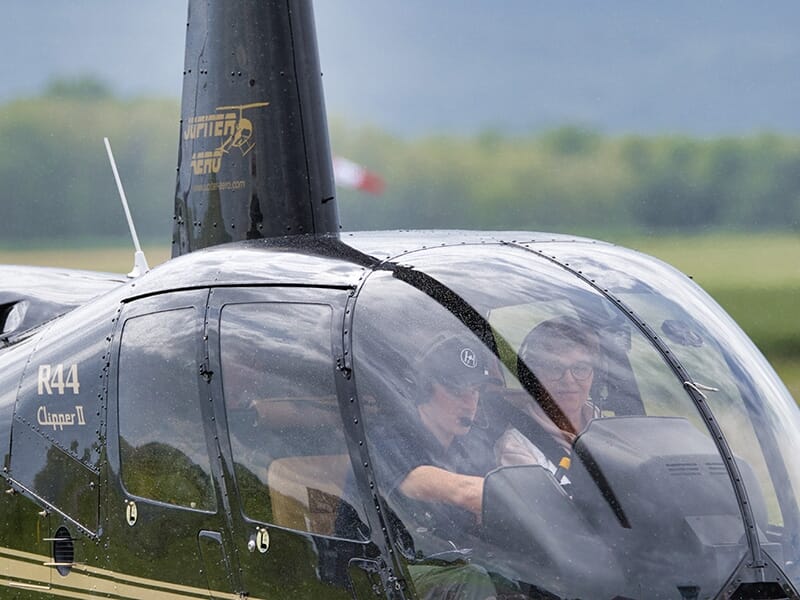 Gros plan sur un hélicoptère noir avec pilote et passager à l'intérieur, prêt au décollage sous un ciel nuageux.