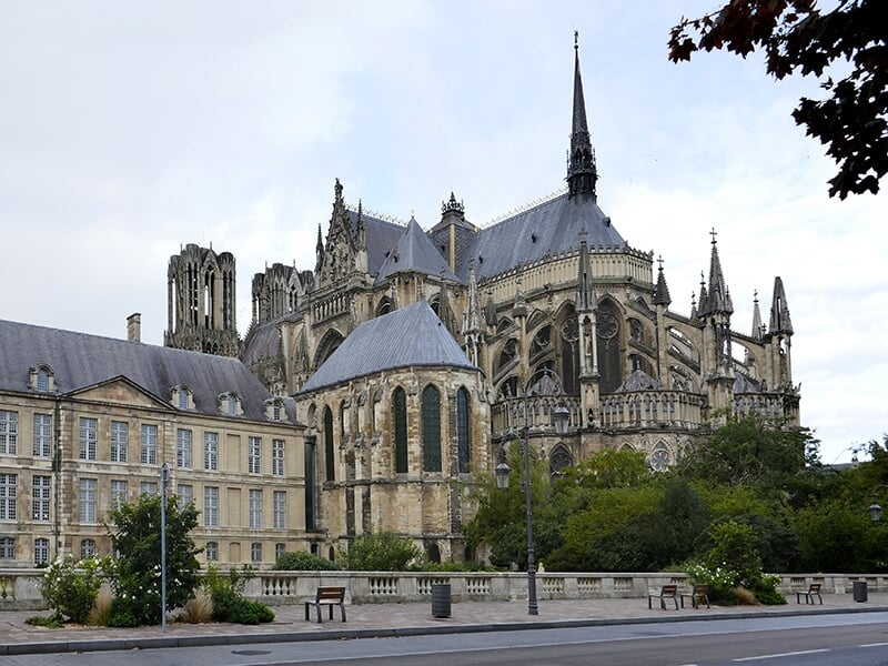 Vue extérieure d'une cathédrale gothique avec des flèches, des arcs-boutants et des vitraux, entourée de bâtiments historiques et d'une végétation luxuriante, prise sous un ciel nuageux.