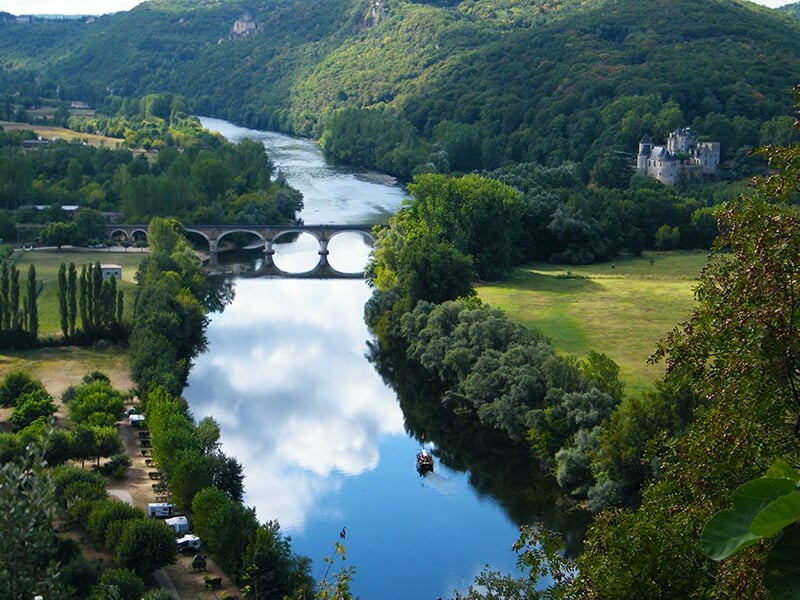Paysage de la Dordogne, montrant une rivière calme entourée de collines verdoyantes, un pont en pierre et un château au loin.
