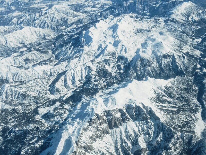 Montagnes enneigées vues d'en haut, avec crêtes abruptes, vallées profondes et forêts sombres parsemées.
