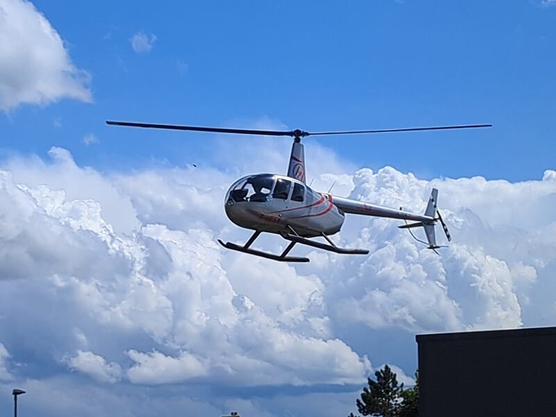 Hélicoptère Robinson R44 volant sous un ciel bleu avec des nuages.