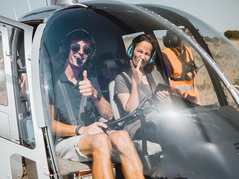 Deux passagers assis à bord d'un hélicoptère, souriants et prêts pour une expérience aérienne. Le pilote et l'un des passagers affichent un geste de satisfaction, probablement avant le décollage.