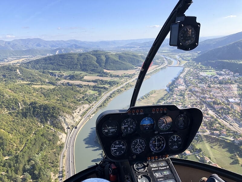 Vue aérienne spectaculaire depuis le cockpit d'un hélicoptère. On peut voir les instruments de bord et un paysage magnifique comprenant une rivière serpentant à travers des collines et une zone habité