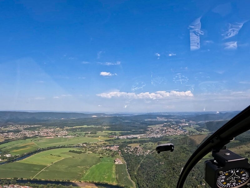 Paysage vallonné avec des champs cultivés, des villages et une rivière serpentant à travers la vallée, vu depuis un hélicoptère. Ciel clair avec quelques nuages blancs, ajoutant de la profondeur à la