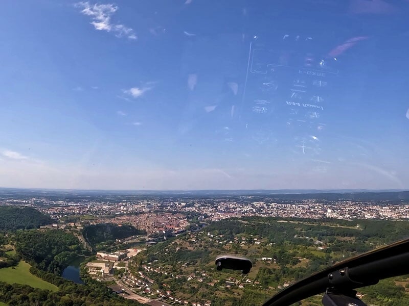Vue aérienne d'une ville entourée de collines verdoyantes, avec un cours d'eau sinueux visible en bas, sous un ciel bleu dégagé. Les reflets du tableau de bord de l’hélicoptère sont perceptibles sur l