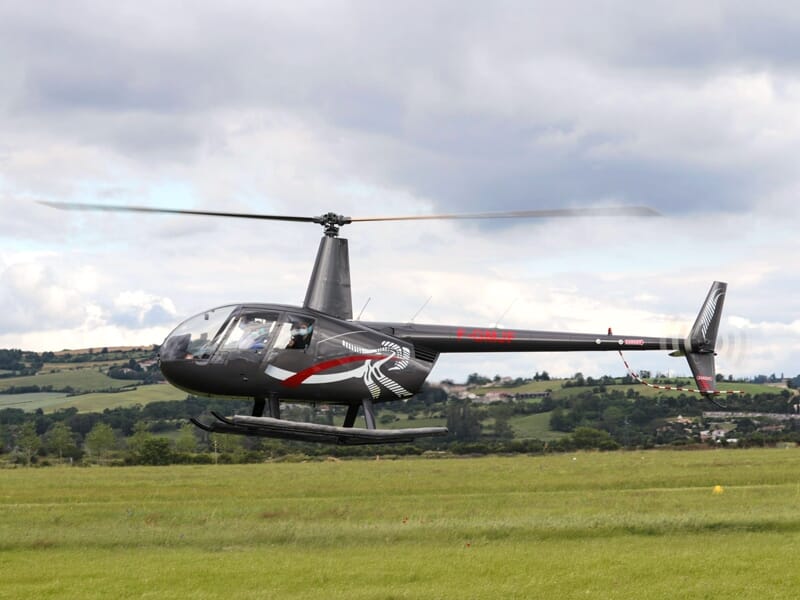 Hélicoptère noir avec des motifs rouges en vol bas au-dessus d'un champ vert avec des collines en arrière-plan.
