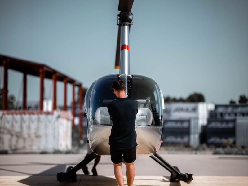 Un homme debout devant un hélicoptère posé au sol, prêt à monter ou inspecter l'appareil.