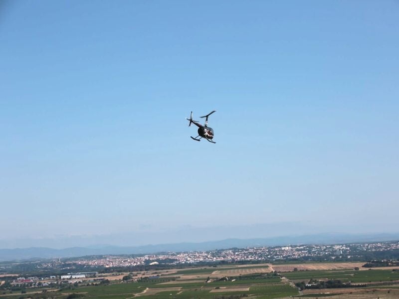Un hélicoptère noir en vol au-dessus d'un paysage rural avec champs et collines sous un ciel bleu clair.
