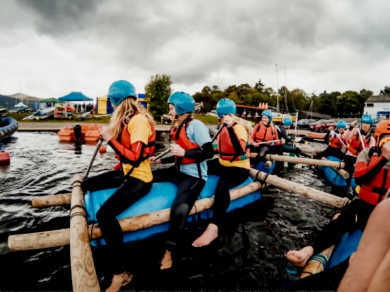 Groupe de personnes en gilets de sauvetage bleus sur un radeau en bois flottant sur un plan d'eau.