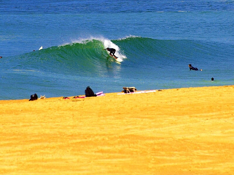 Surfeur attrapant une vague bleue sur une plage de sable doré à Soorts-Hossegor sous un ciel clair.