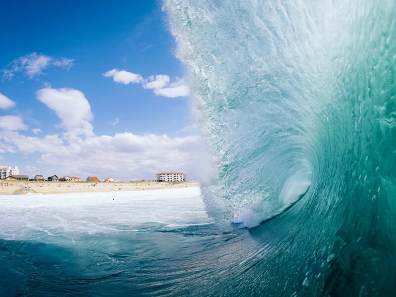 Vague puissante formant un tube bleu turquoise avec un ciel partiellement nuageux en arrière-plan.