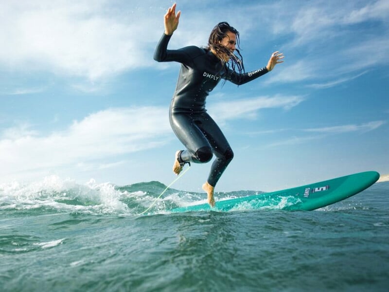 Surfeuse en combinaison noire debout sur sa planche, glissant sur une vague avec un ciel bleu clair.