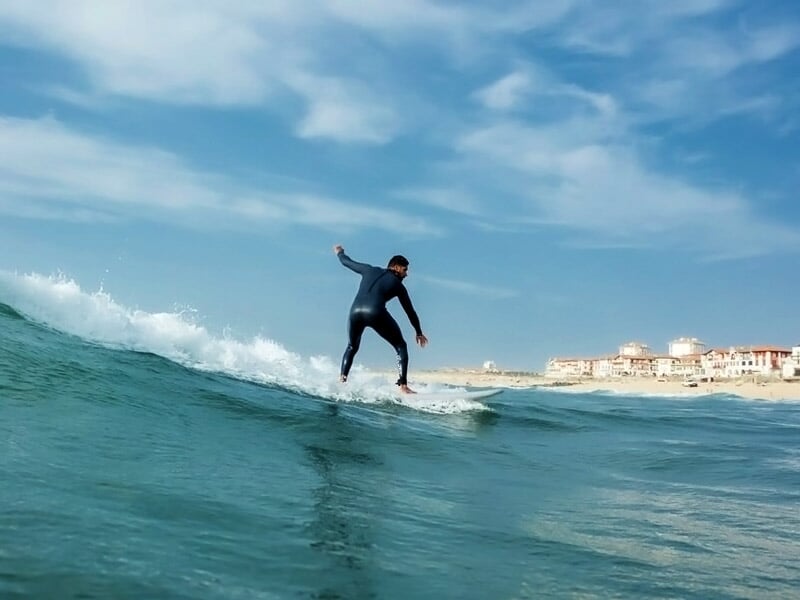 Surfeur en combinaison noire surfant une vague calme près de la côte avec des bâtiments en arrière-plan.