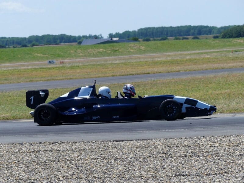 Voiture de Formule 3 bleu foncé sur une piste de course, avec deux conducteurs casqués en pleine vitesse sur une piste entourée de verdure.