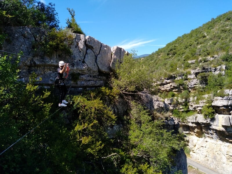 Personne en via ferrata suspendue à une corde au-dessus d'une vallée rocheuse et boisée sous un ciel bleu clair.
