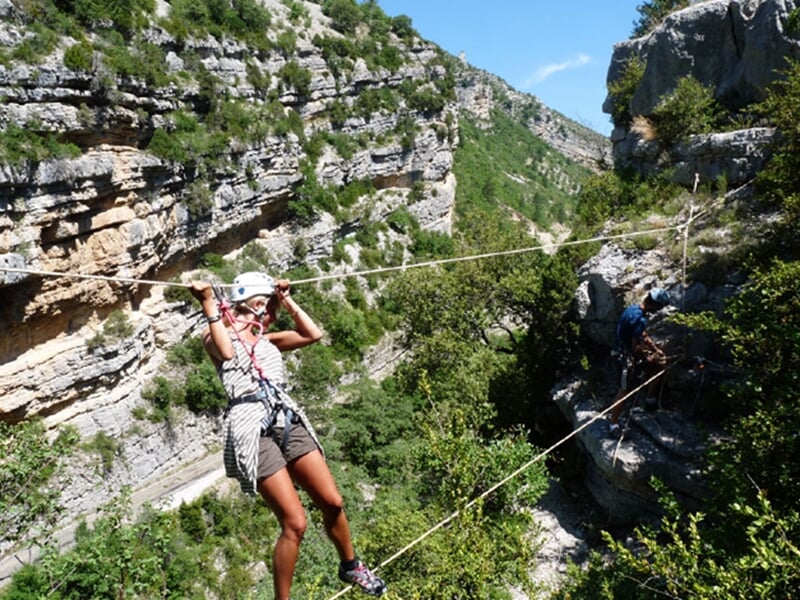 Femme équipée d'un casque et harnais traverse une via ferrata entre des falaises rocheuses et une végétation dense.