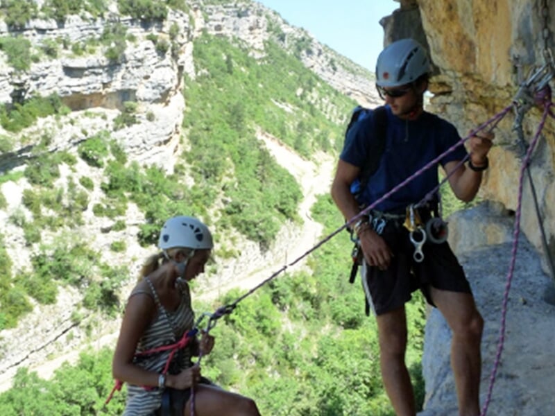 Deux personnes équipées pour l'escalade discutent sur une paroi rocheuse avec un paysage de vallée en arrière-plan.