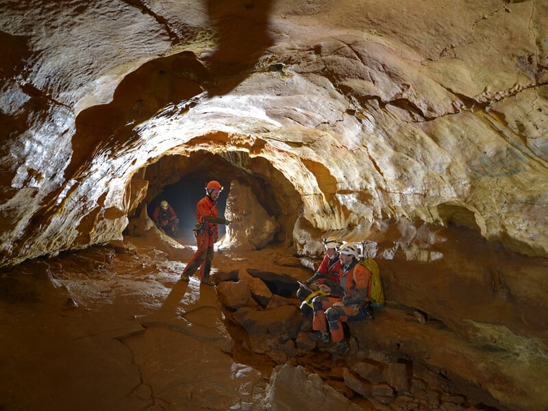Groupe de spéléologues en tenue rouge et casque dans une grande salle souterraine rocheuse avec des équipements.