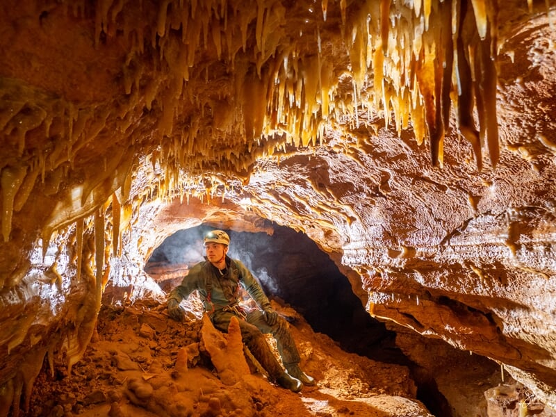 Spéléologue assis dans une grotte ornée de nombreuses stalactites lumineuses et de formations rocheuses.