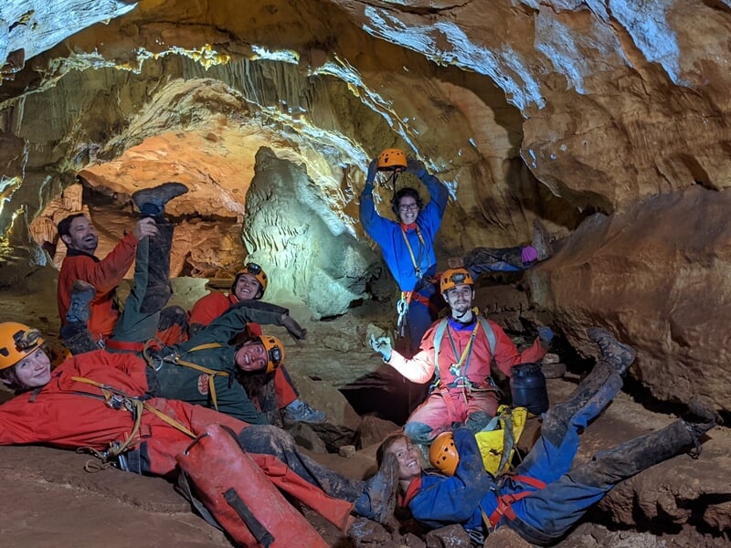 Groupe de spéléologues en combinaison rouge posant joyeusement dans une grande cavité rocheuse souterraine.
