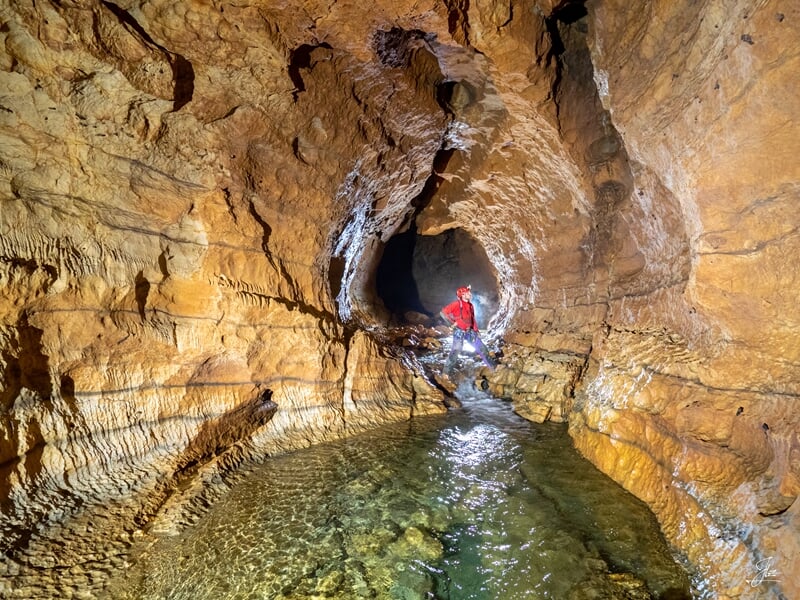Spéléologue en combinaison rouge traverse une rivière souterraine dans une galerie rocheuse éclairée.