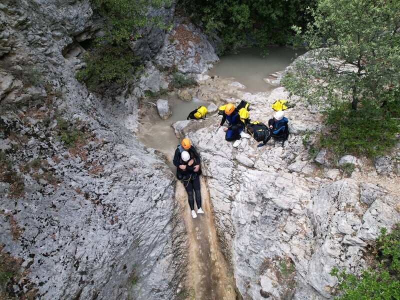 Personne équipée descend en rappel sur une paroi rocheuse dans un canyon avec un groupe à proximité.