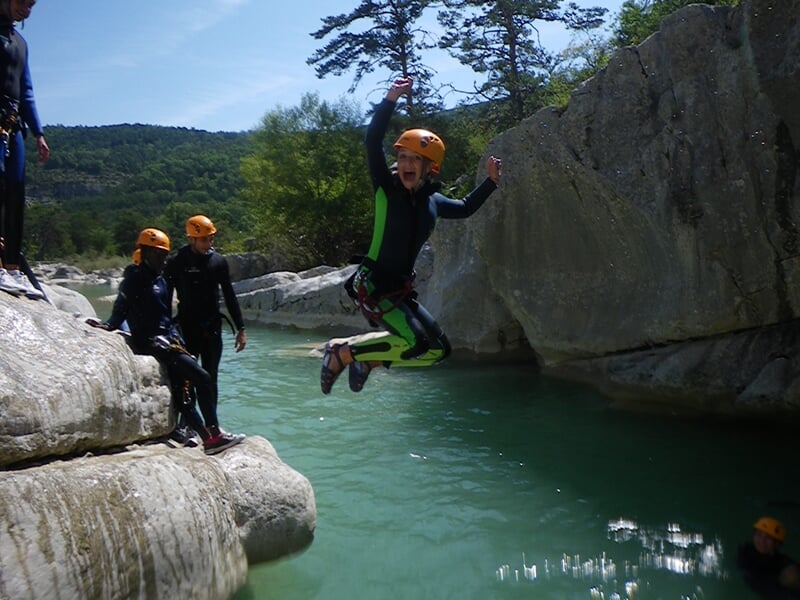 Personne en combinaison noire glisse sur un toboggan naturel rocheux dans un canyon entouré de végétation.