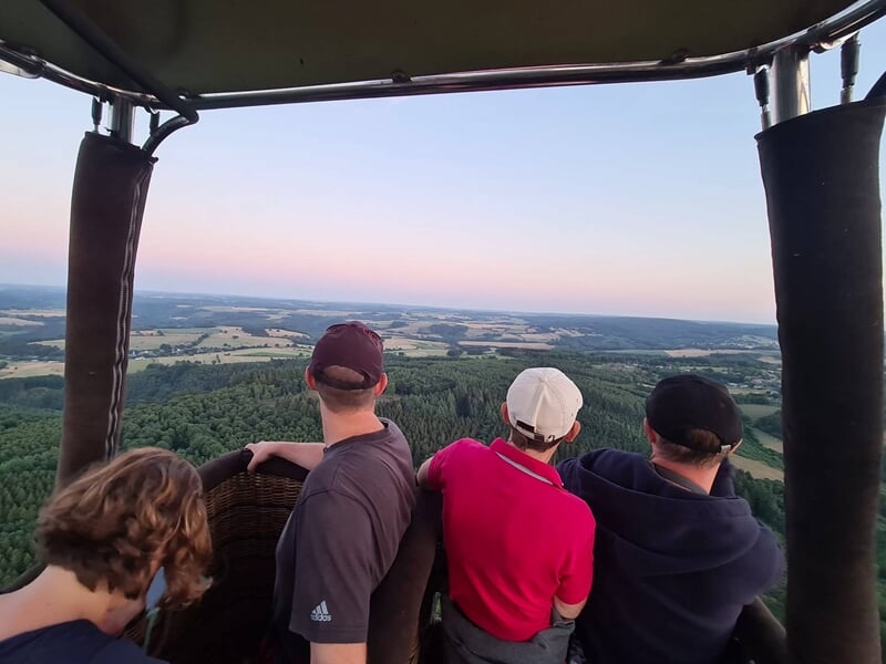 Quatre personnes dans une nacelle de montgolfière survolant une vaste campagne au coucher du soleil.