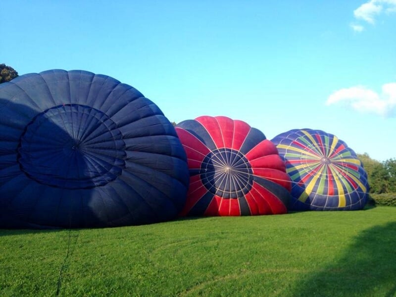 Trois montgolfières dégonflées posées sur une pelouse verte sous un ciel bleu clair.